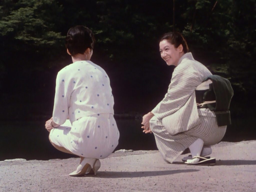 still from The End of Summer (1961) - two Japanese women, one in a dress and high heels, one in a kimono and sandals, crouch on sand next to a river